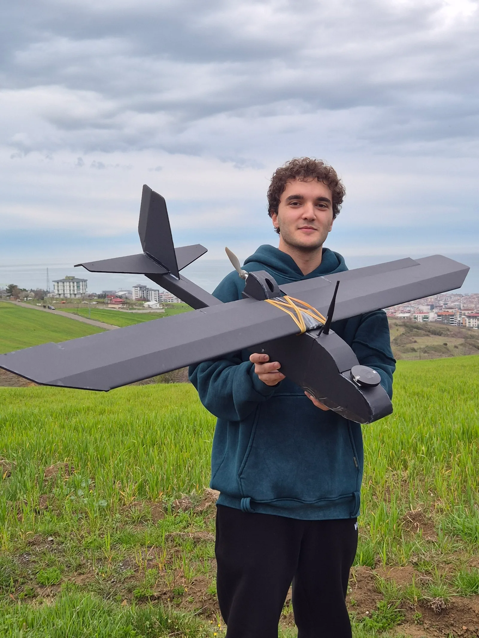 A young man on a green hill holding the black model plane he built